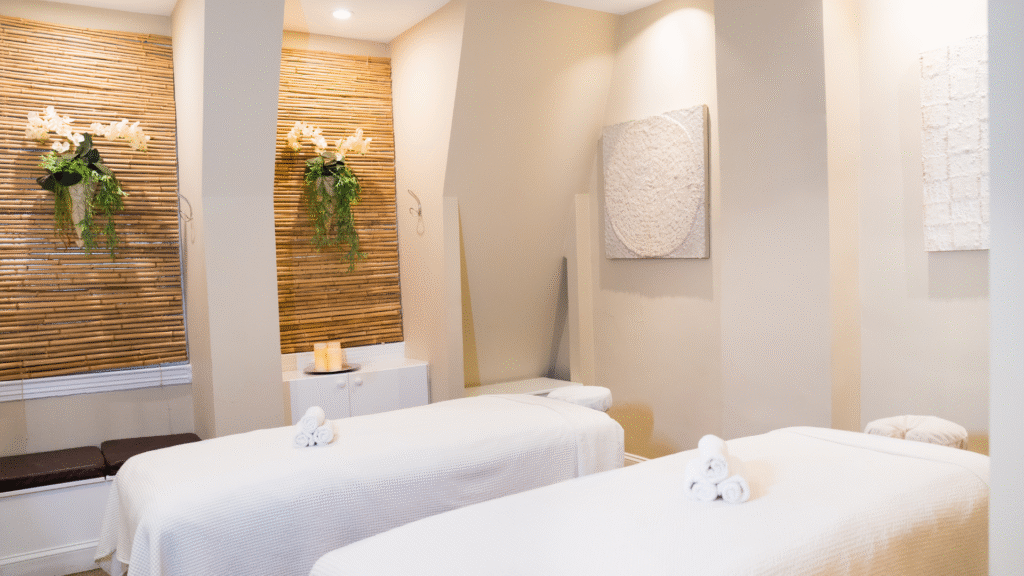 Two massage tables neatly arranged and clean. Bamboo and white flowers on the walls, and three lit candles on top of the cabinet. A very cozy and welcoming atmosphere.
